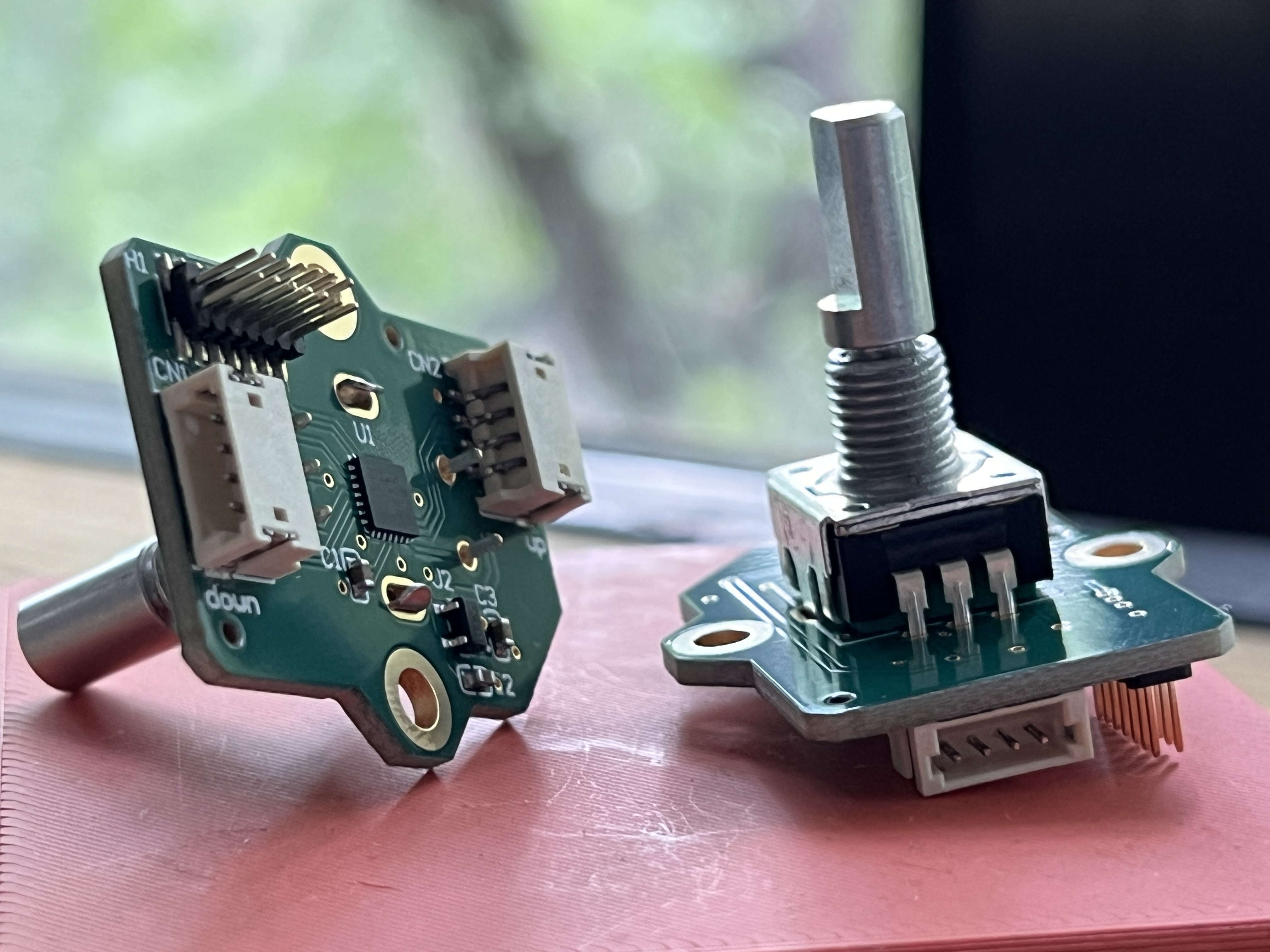 two PCBs sit next to each other. they are "buoys", distributed serial control nodes. one is on its side showing the circuitry underneath, one sitting flat with the encoder, which is opposite the circuitry, pointing upwards. they sit on a small pink plastic surface with blurry trees through a window behind. the light is kind of dramatic from outside.